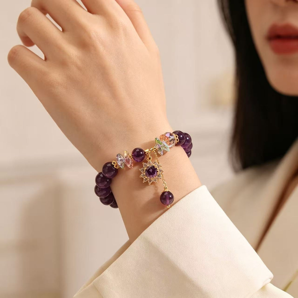 Wrist close-up of an amethyst bracelet with rainbow crystal accents and a gold-tone flower charm, styled with a white sleeve.